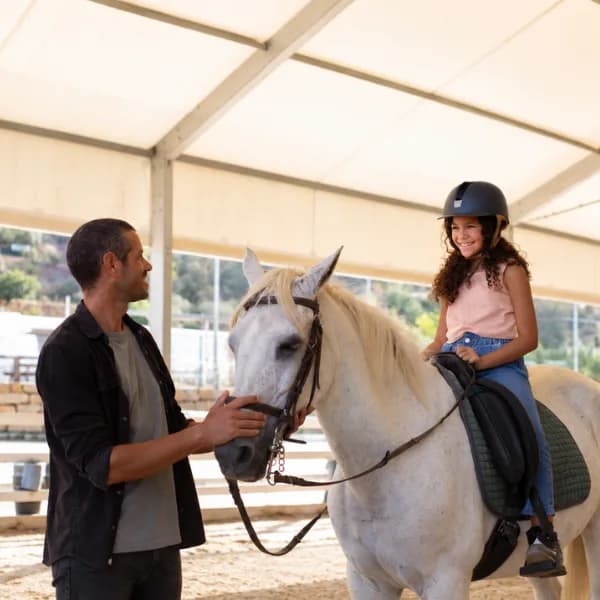 Ein Reitlehrer führt ein Schimmel-Pony, auf dem ein Mädchen mit Reithelm sitzt